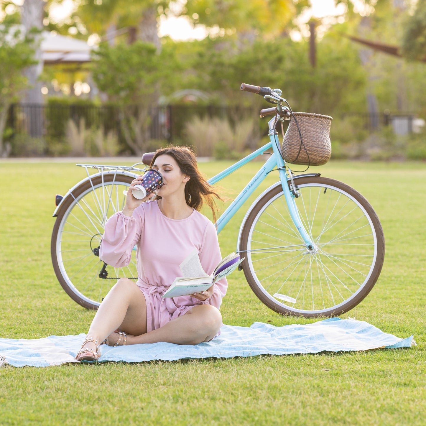 Young lady sitting on a picnic rug reading and drinking coffee with her bike behind her and a warners wheels bicycle basket attached